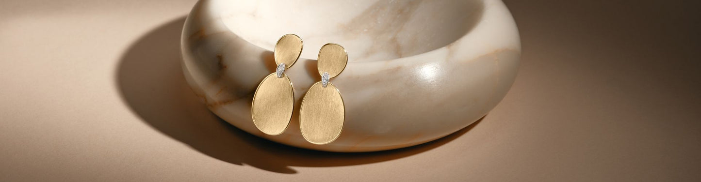 Pair of modern gold drop earrings with brushed oval shapes and small diamond accents, displayed against a sculptural marble background.