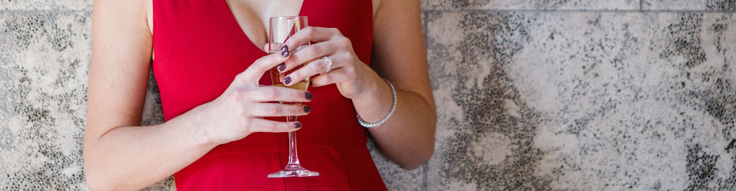 Woman in a red dress holding a champagne flute, wearing a diamond ring and bracelet, styled against a textured stone background.