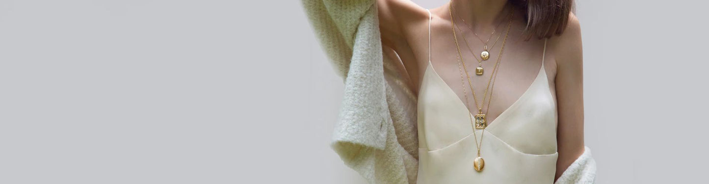 Model wearing layered gold necklaces with delicate pendant charms, styled over a cream silk dress and soft textured wrap for an elegant, minimalist look.
