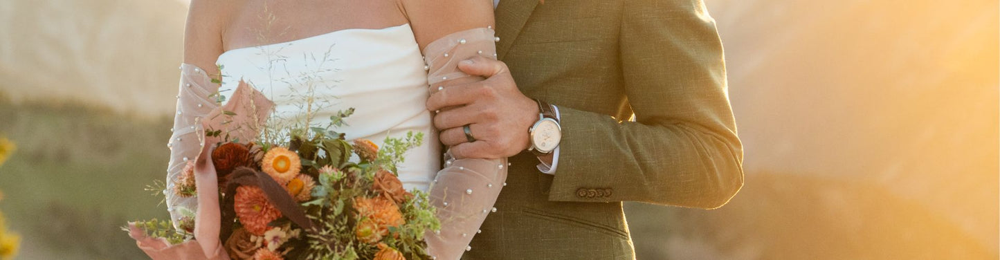 Bride and groom standing together at sunset, with the groom wearing a wedding band and watch while holding the bride’s arm as she carries a floral bouquet.